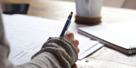 A hand holding a pen and writing on paper upon a wooden desk.